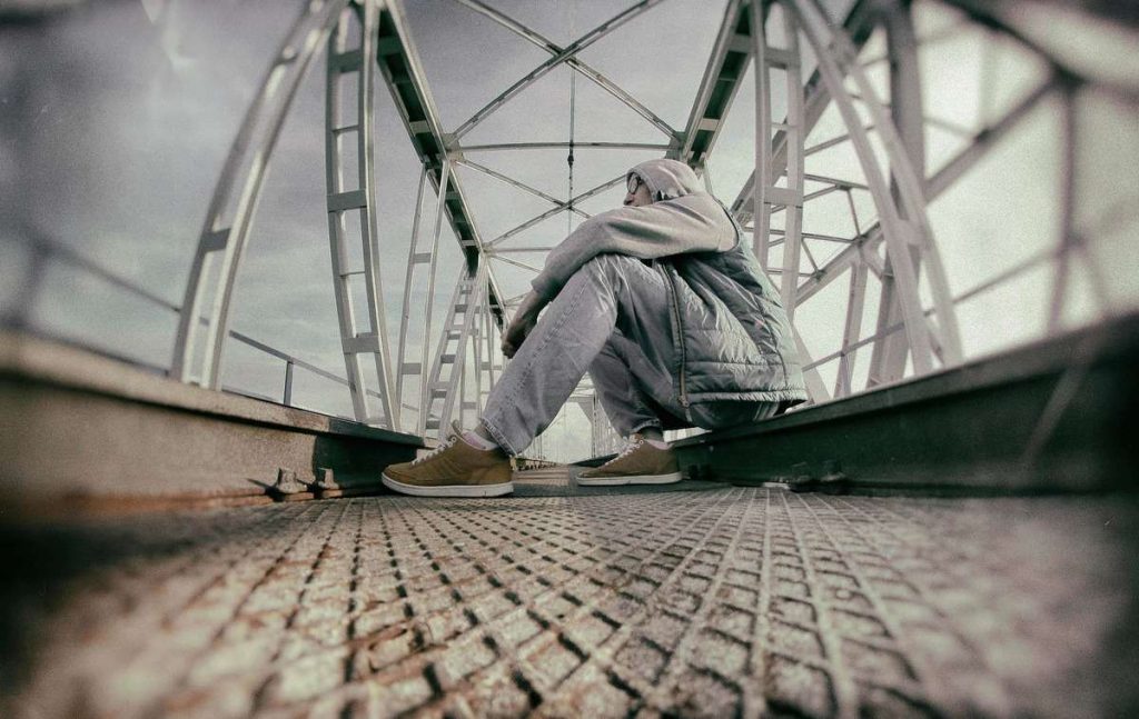 Persona encapuchada sentada sola en un puente metálico con la cabeza baja