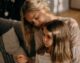 Madre abrazando a su hija pequeña en el sofá mientras la niña mira hacia abajo con expresión triste