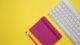 Cuaderno rosa con un lápiz junto a un teclado blanco sobre un fondo amarillo
