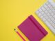 Cuaderno rosa con un lápiz junto a un teclado blanco sobre un fondo amarillo