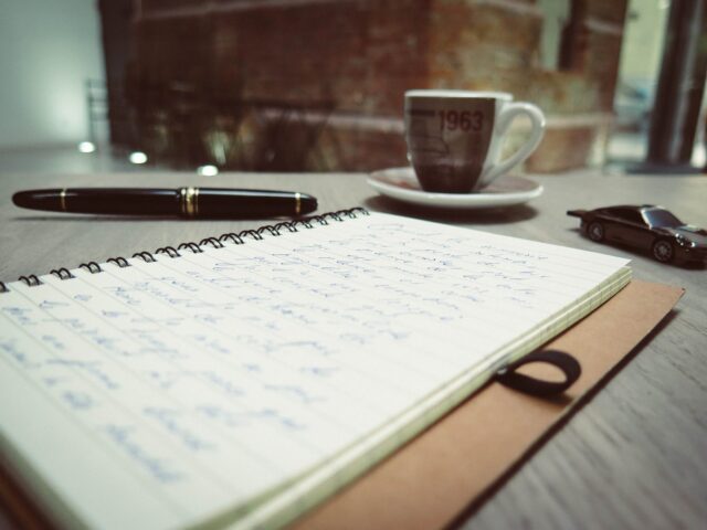 closeup photo of blue pen tinted spiral notepad placed beside pen die cast car and coffee cup
