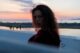 Mujer joven de pelo largo y rizado que mira de reojo a la cámara frente al mar al atardecer