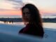Mujer joven de pelo largo y rizado que mira de reojo a la cámara frente al mar al atardecer
