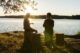 Dos personas conversan sentadas en troncos a la orilla de un lago, a contraluz del sol del atardecer