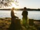 Dos personas conversan sentadas en troncos a la orilla de un lago, a contraluz del sol del atardecer