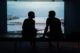 Siluetas de dos personas sentadas frente a un gran ventanal con vistas al mar, conversando