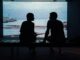 Siluetas de dos personas sentadas frente a un gran ventanal con vistas al mar, conversando