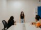 Una mujer de pie habla frente a un grupo de personas sentadas alrededor de una mesa en una sala blanca