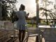 Mujer joven con vestido blanco de pie en una terraza de madera mirando su móvil al atardecer