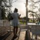 Mujer joven con vestido blanco de pie en una terraza de madera mirando su móvil al atardecer