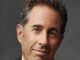Retrato del comediante Jerry Seinfeld sonriendo con chaqueta oscura sobre fondo gris