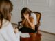 Joven sentada en un sillón llorando con la cara entre las manos frente a una terapeuta de espaldas