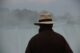 Hombre de espaldas con abrigo oscuro y sombrero frente a un paisaje con niebla