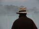 Hombre de espaldas con abrigo oscuro y sombrero frente a un paisaje con niebla