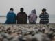 Cuatro adolescentes sentados de espaldas en una playa de piedras mirando el mar