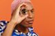 Hombre de pelo rosa que mira a través de una lupa redonda sostenida frente a su ojo, sobre fondo naranja