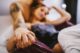 Pareja en la cama tomada de la mano en primer plano, ella apoyada en el hombro tatuado de él