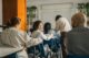 Estudiantes sentados en pupitres en un aula mientras un profesor se inclina a ayudar a una alumna