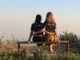 Dos hermanas de espaldas, sentadas en un banco al atardecer y abrazadas, con camisetas que dicen «Sisterhood»; imagen ilustrativa sobre la esquizofrenia en hermanos.